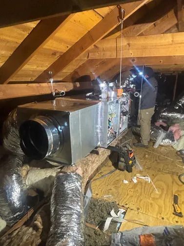 Attic HVAC unit with silver ductwork under wooden rafters, lit by a bright work light.
