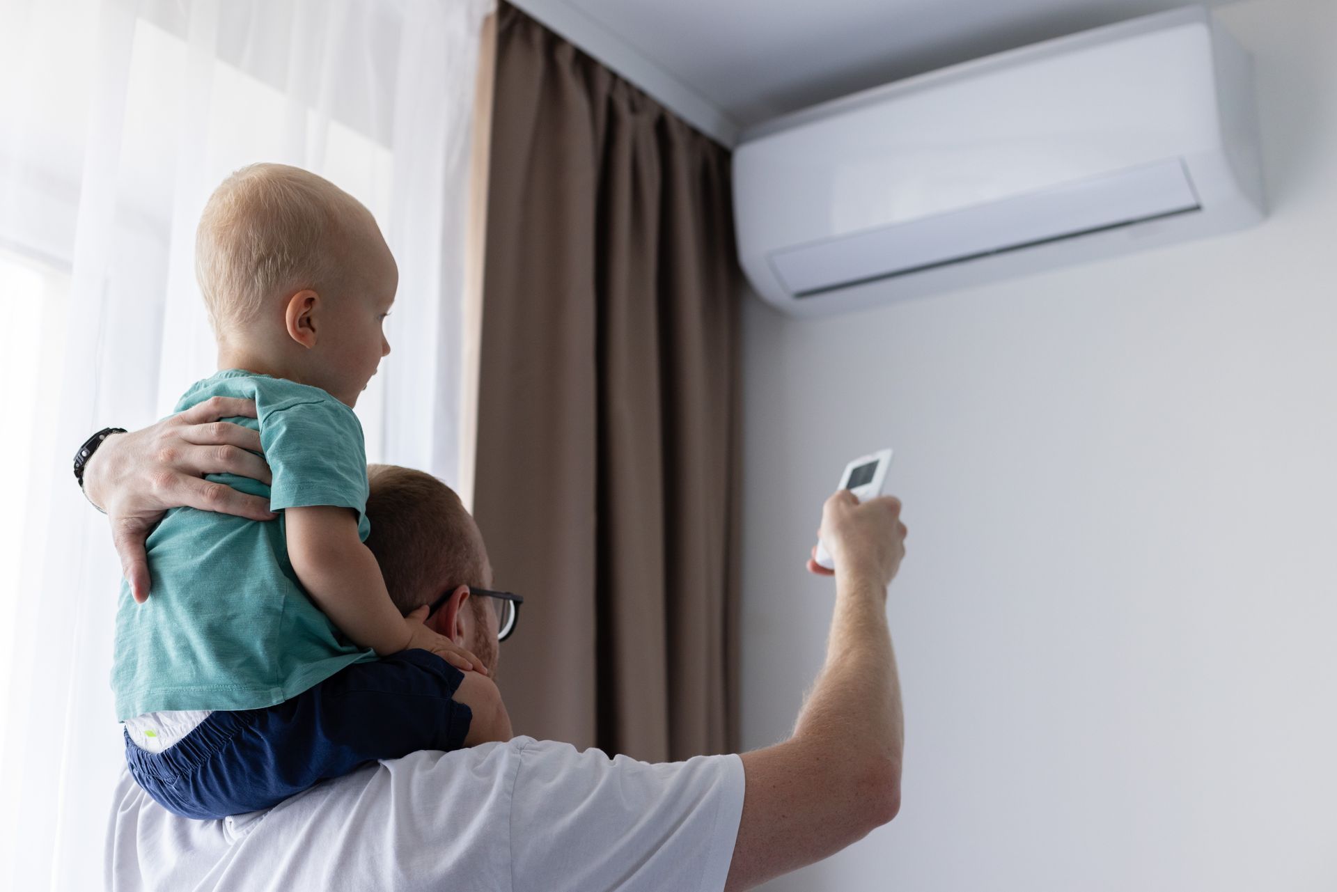 A person holding a child on their shoulders uses a remote control to adjust an air conditioner unit mounted on a wall.