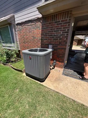 Outdoor air conditioner unit beside a brick house wall and window on a sunny lawn path