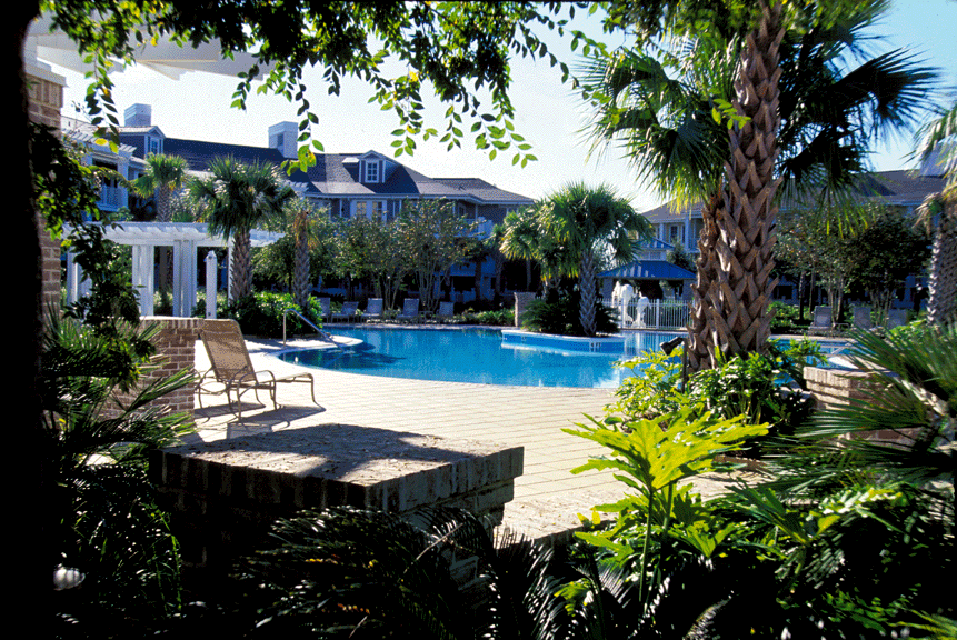 Swimming pool flanked by trees and small stone buildings under a wooden pergola.