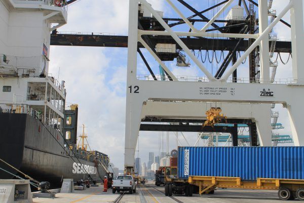 Una grúa portuaria carga un contenedor marítimo azul en un camión junto a un buque de carga en un muelle concurrido
