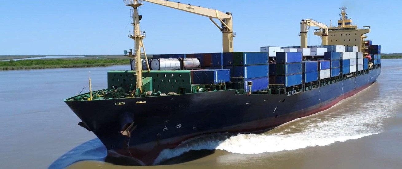 Buque de carga con contenedores azules navegando por un río cerca de la orilla