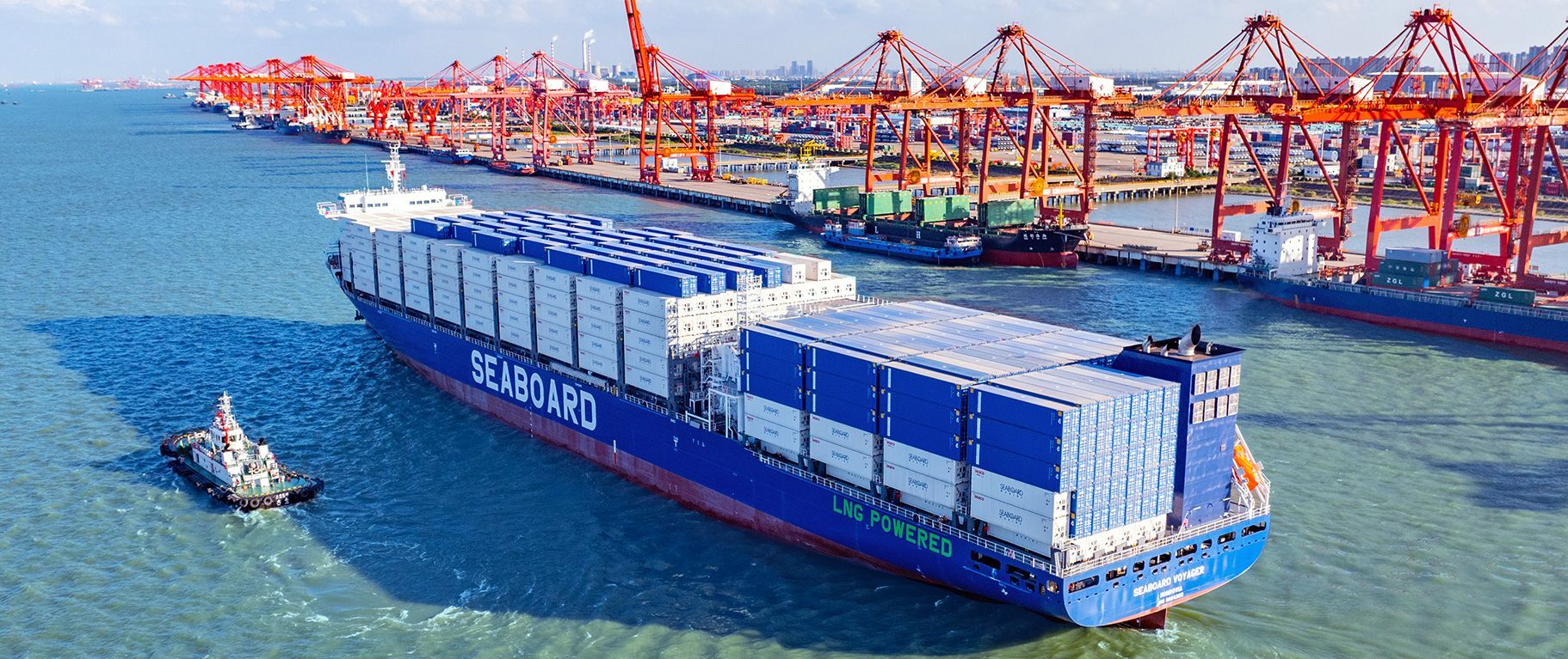 Buque de carga azul en el agua, cerca de un puerto de contenedores concurrido con grúas rojas