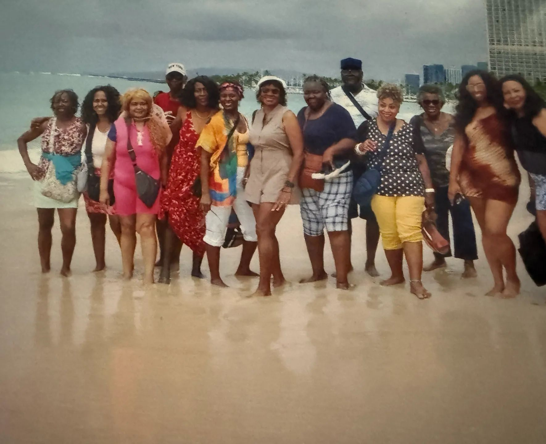 Group of people standing barefoot on a beach, smiling and posing in colorful clothes by the ocean.