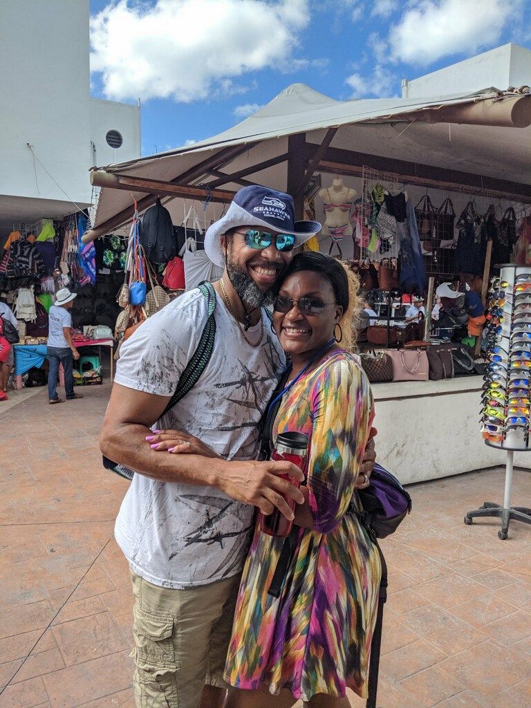 Couple hugging, smiling in front of outdoor market with souvenirs and blue sky.