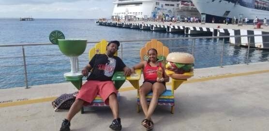 Two people sit in novelty chairs by a cruise ship. The chairs are margarita and burger themed.