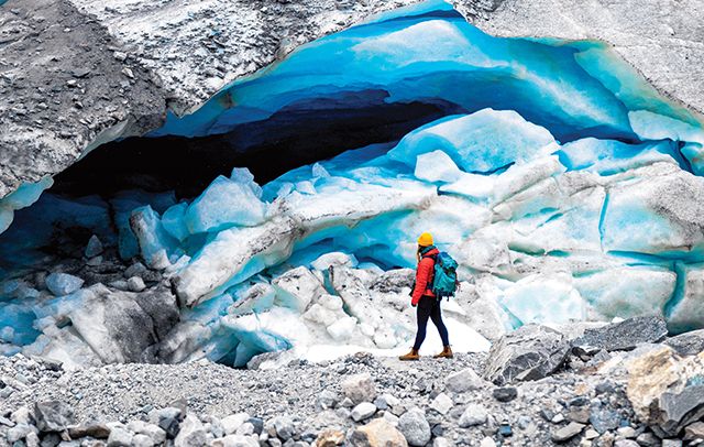 Woman in orange jacket and yellow helmet near a blue glacial cave.