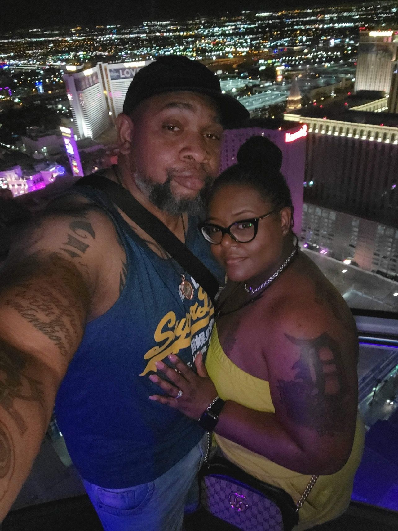 Couple in Las Vegas at night, cityscape backdrop; man with tattoos, woman with glasses.