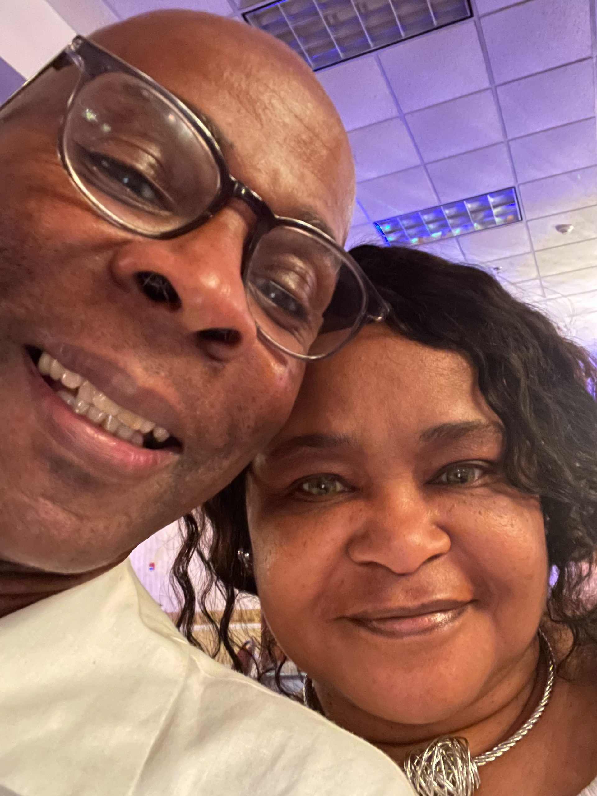 Two smiling people, a man in glasses and a woman, close-up, possibly at an event with a silver necklace.