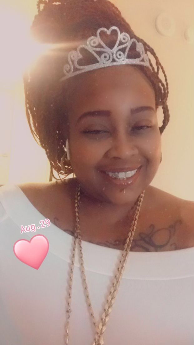 Woman wearing a tiara and gold necklace smiles at the camera; heart emoji to the side.