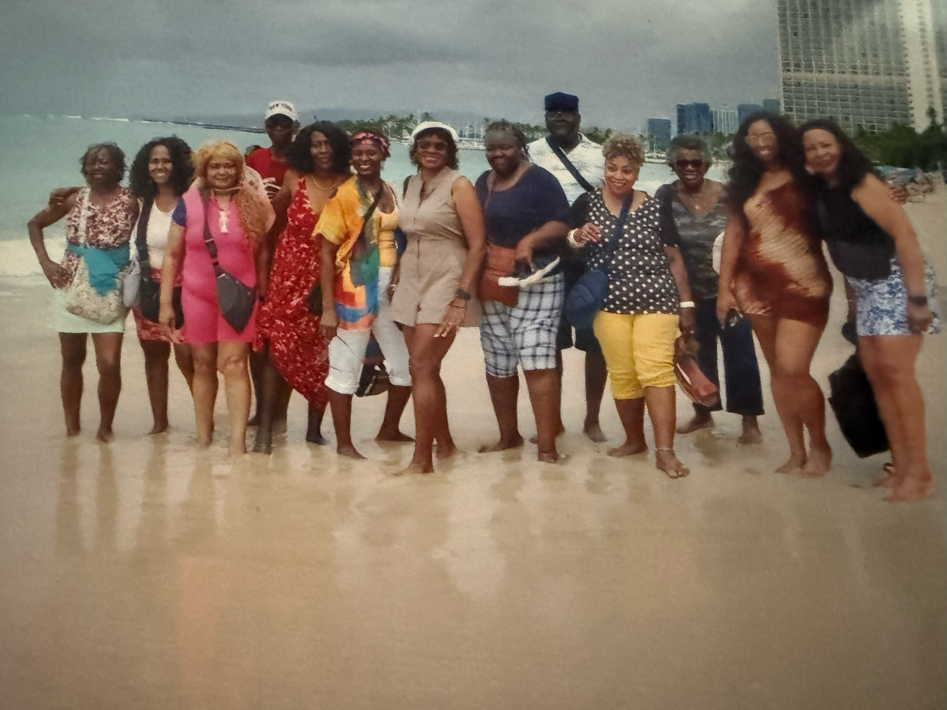 A group of 14 people stand in a line on a beach for a photo, with city buildings visible in the background.
