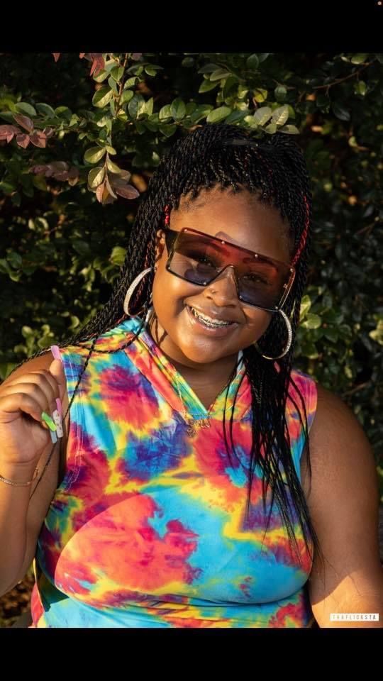 Woman with braids wearing sunglasses and a tie-dye top, smiling.