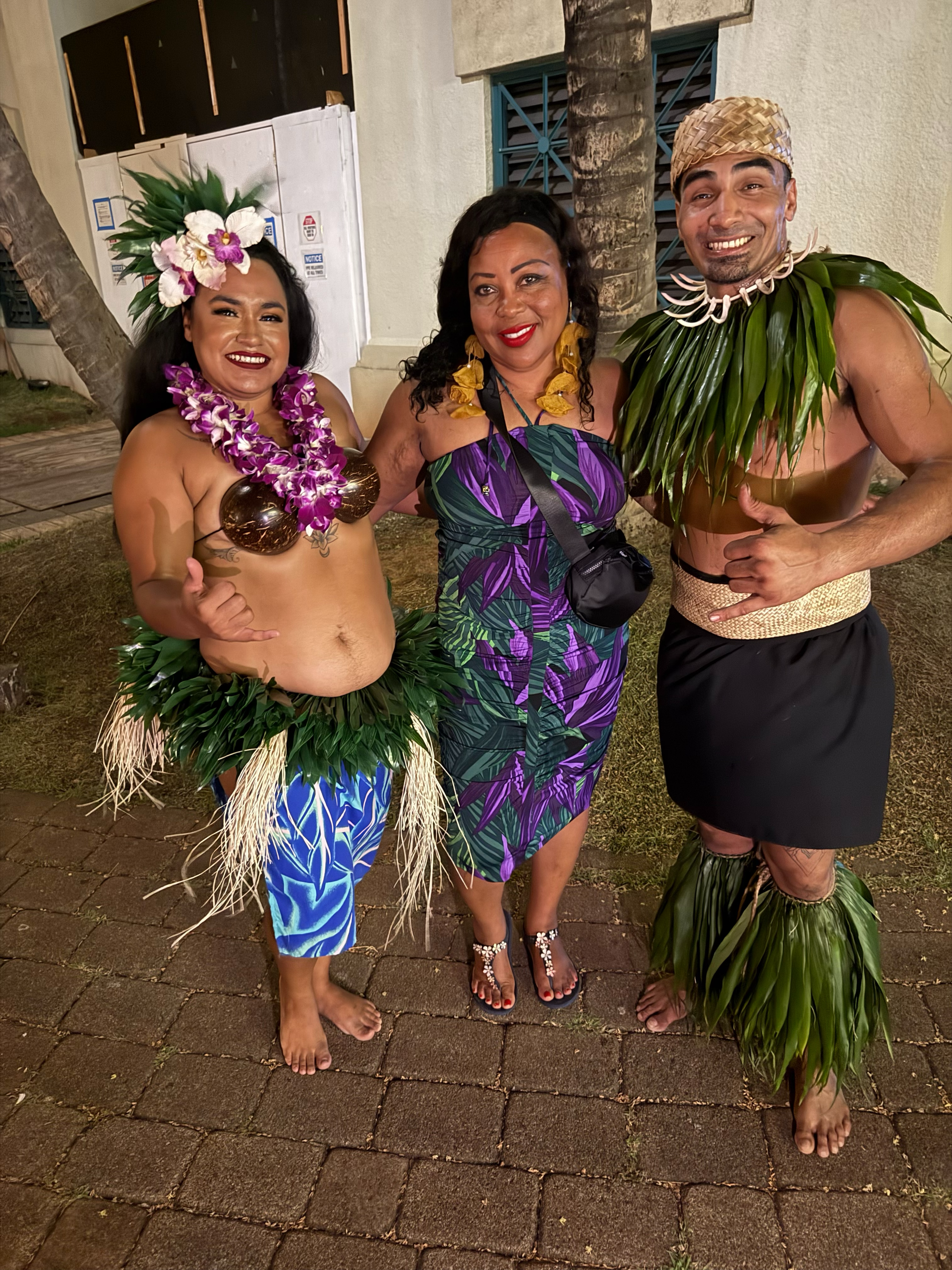 Three people smiling while posing together outdoors, wearing traditional Polynesian attire and a patterned dress.