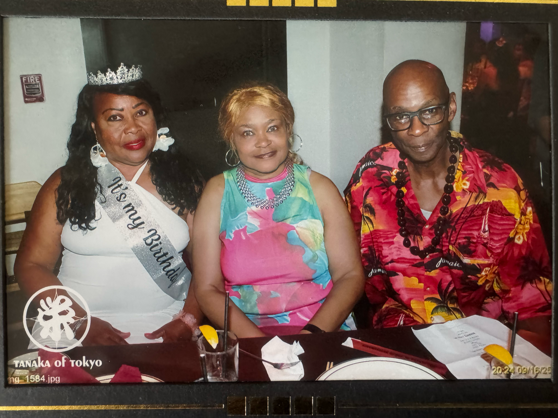 Three people sit at a Tanaka of Tokyo restaurant table; the woman on the left wears a tiara and a birthday sash.