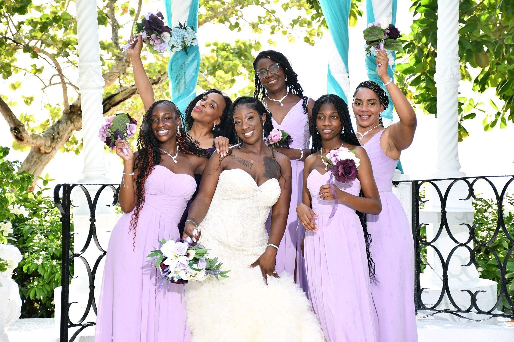 Bride and bridesmaids in lavender dresses pose, smiling, at outdoor wedding.