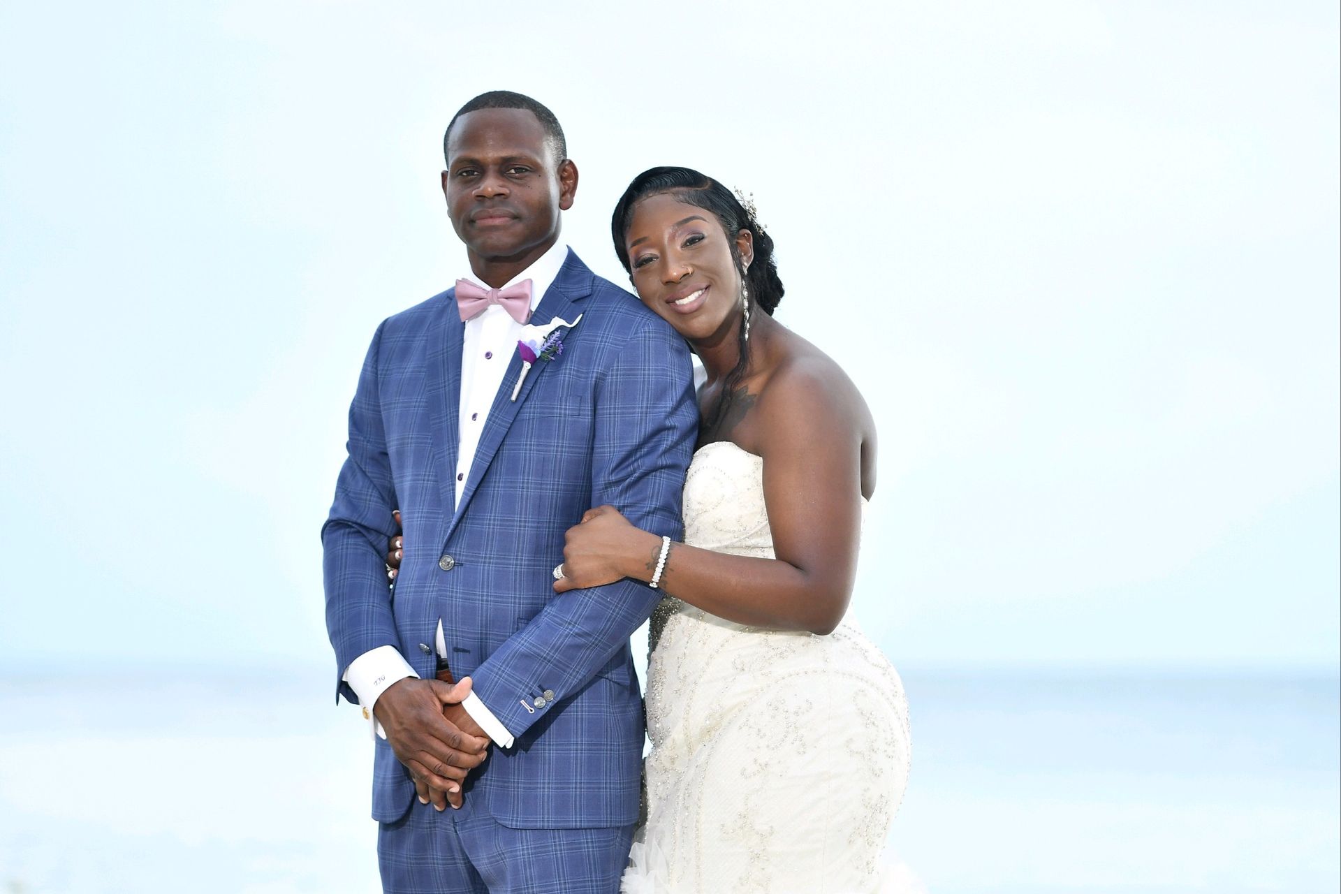 Bride and groom pose together, ocean background. Groom in blue suit, pink bow tie. Bride in strapless white dress.