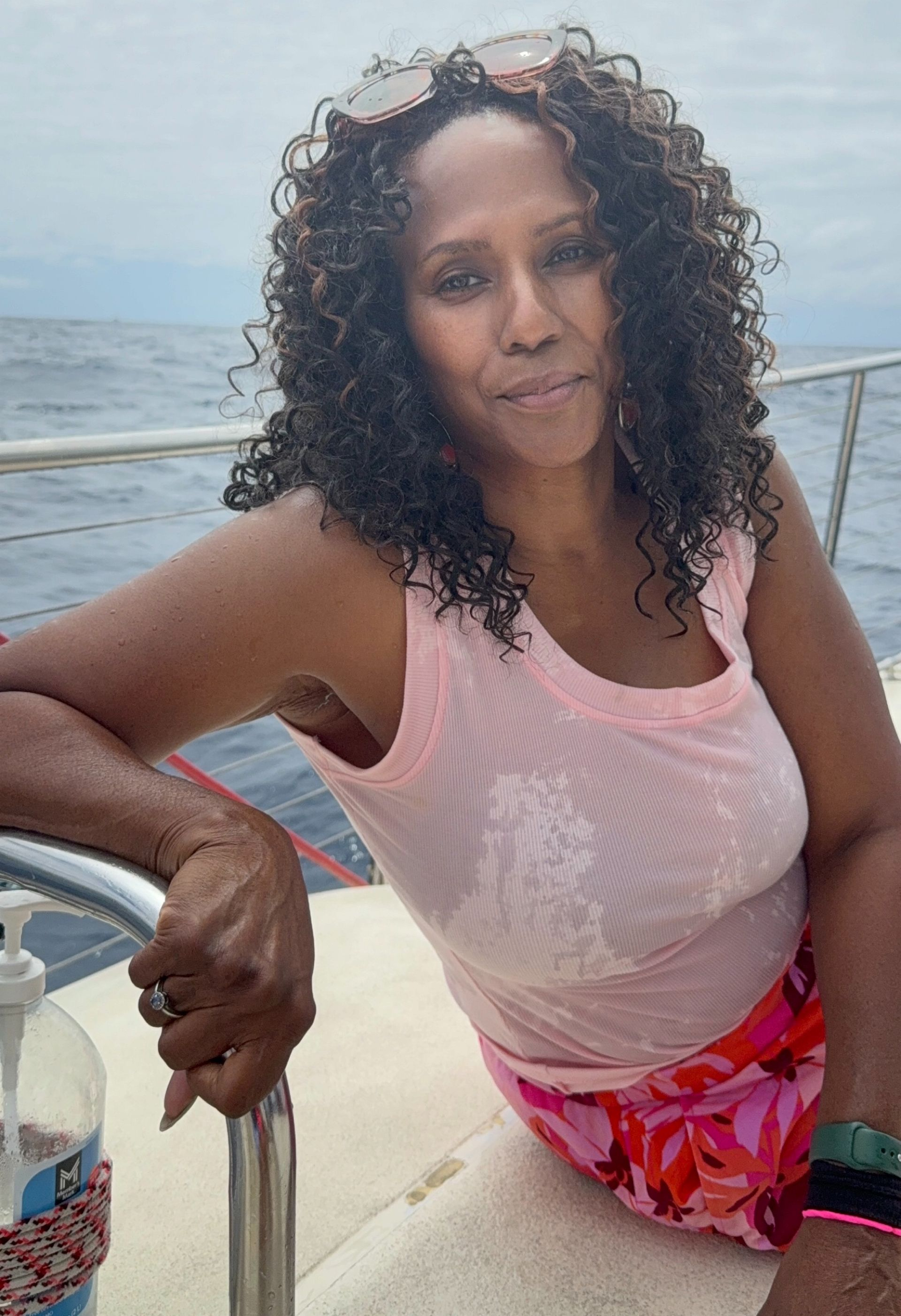 A woman with curly dark hair and a light pink tank top sits on a boat, leaning against a railing with the sea behind her.