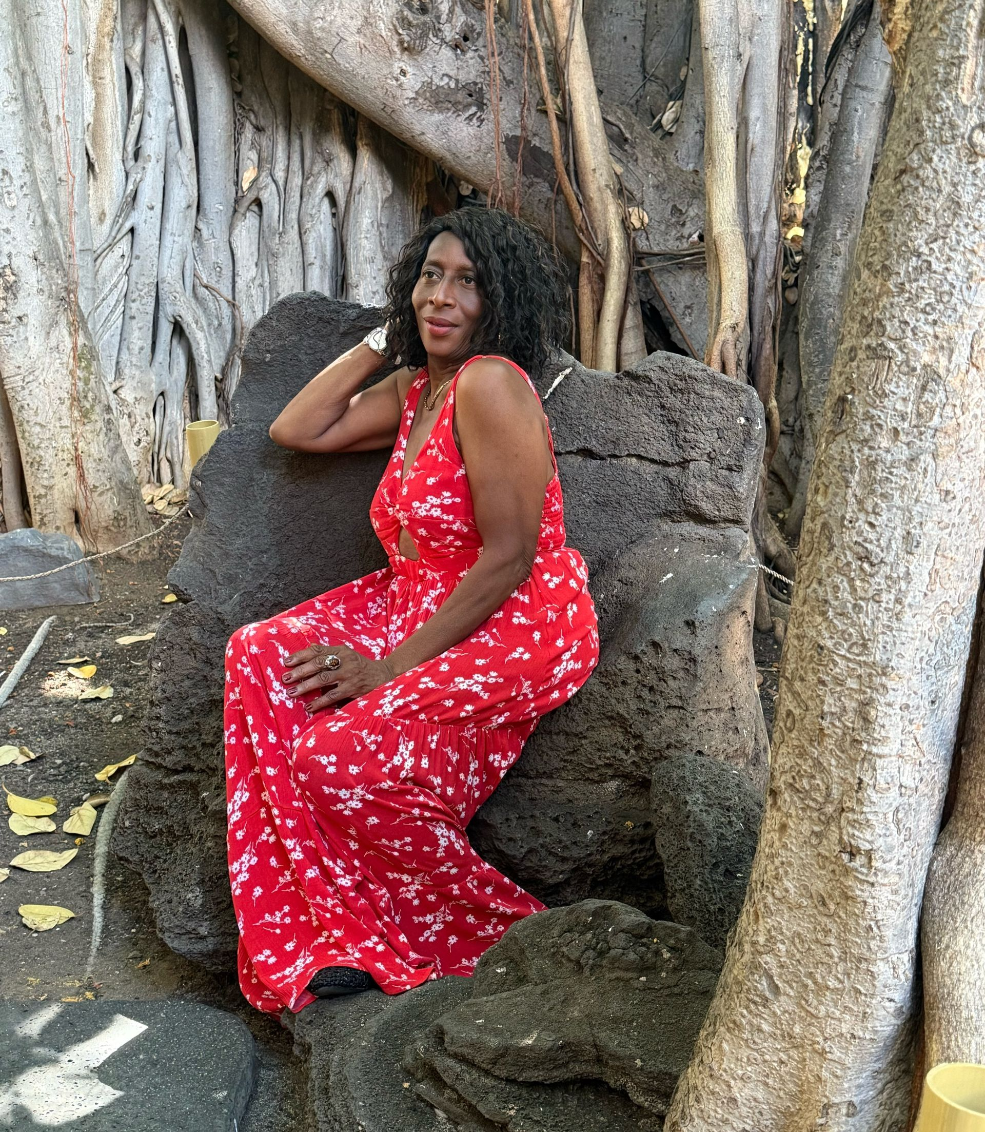 A person in a red floral maxi dress sits on a natural stone seat between the roots of a large banyan tree.