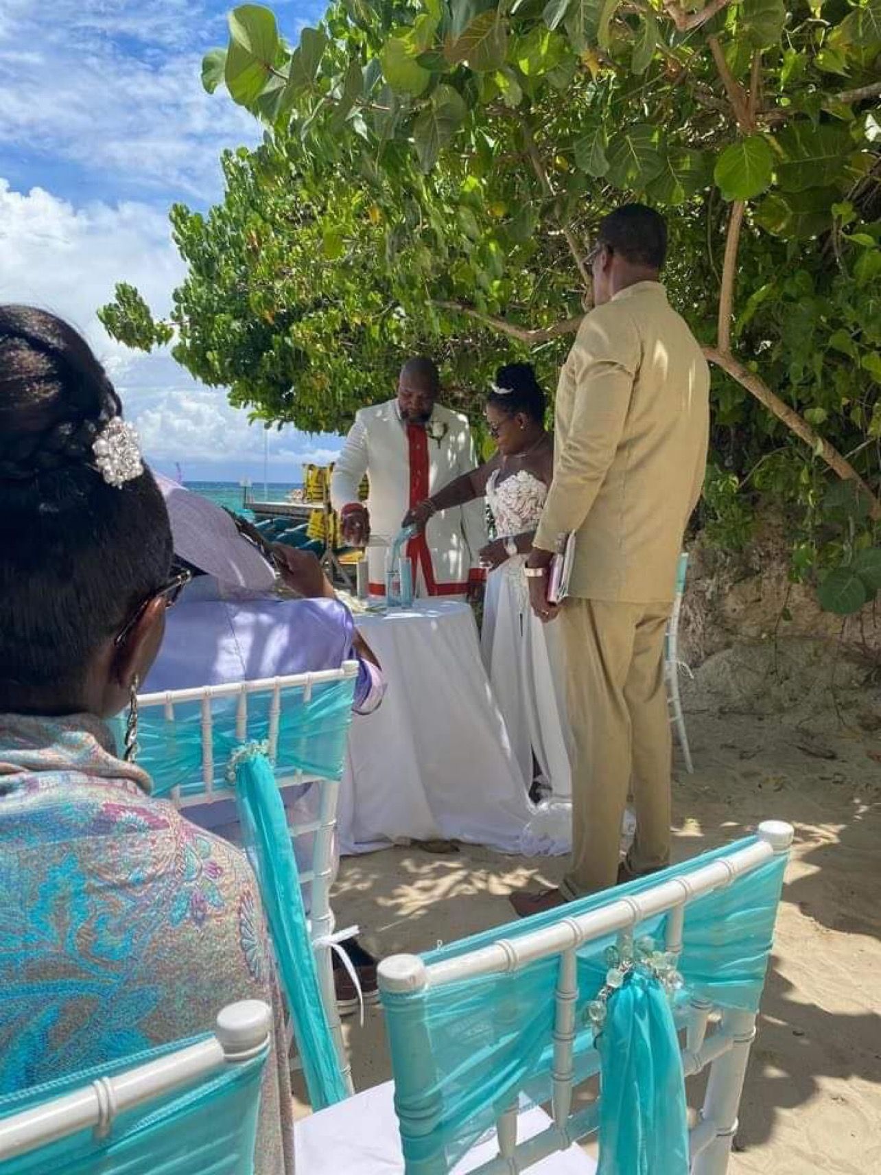 Wedding ceremony on a beach: Bride and groom pour sand. Teal chairs, white tablecloth, sunny day.