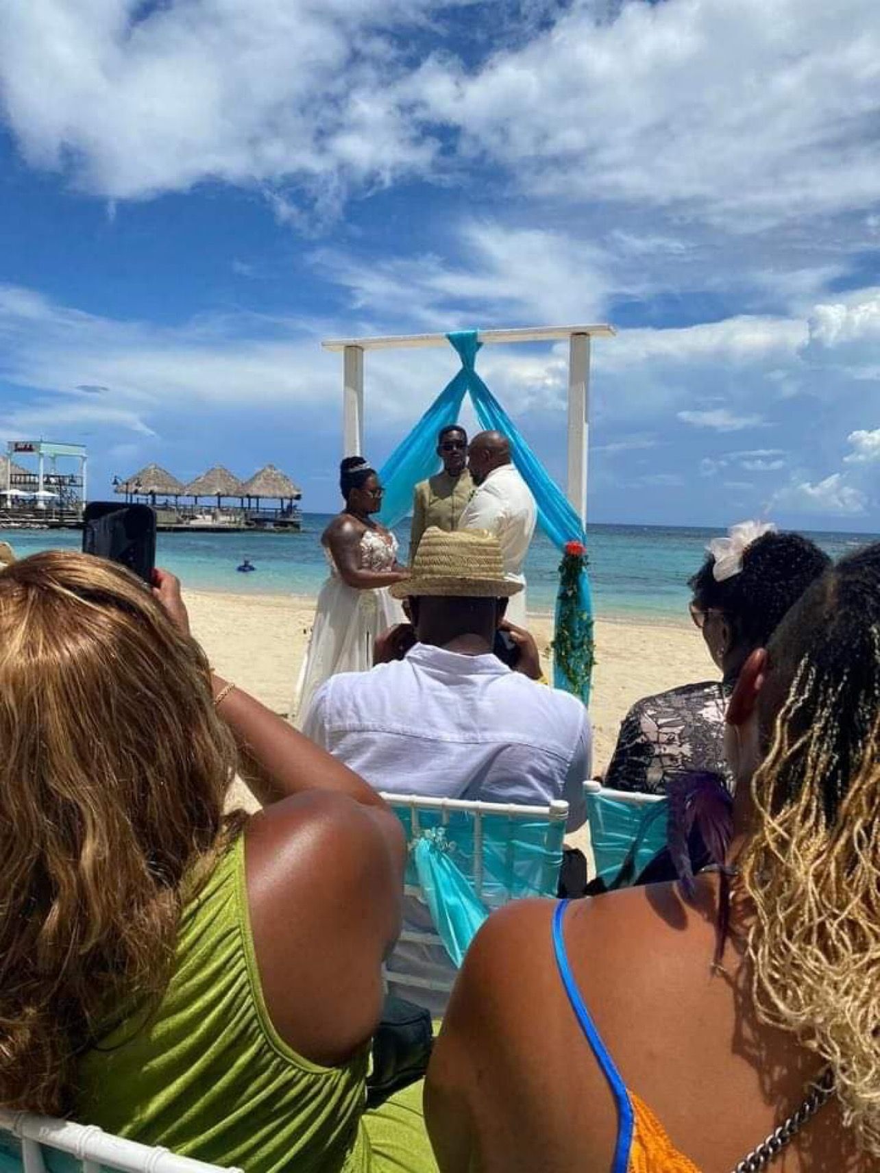 Beach wedding with bride and groom under an arch. Guests watch. Blue sky and ocean in the background.
