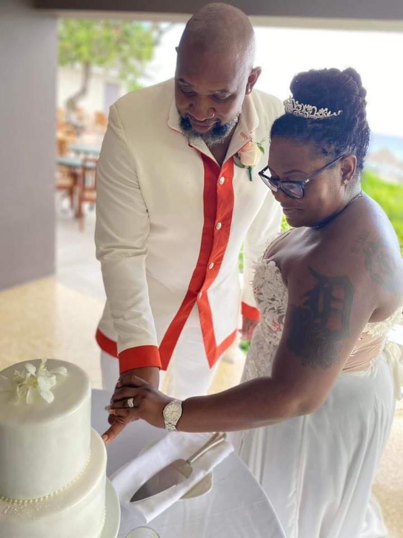 Couple cuts wedding cake: Man in white suit, woman in white dress with tiara, outdoor setting.