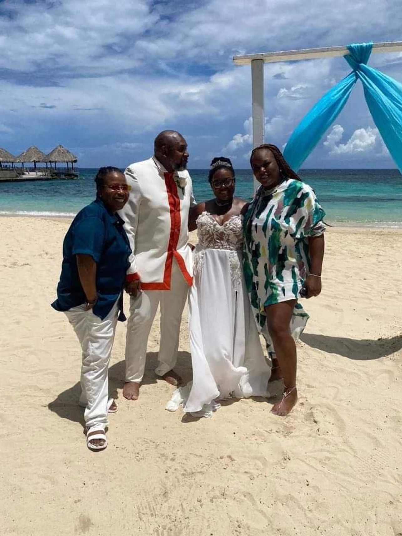 Wedding group on beach with bride in white gown and groom in white suit; ocean in background.