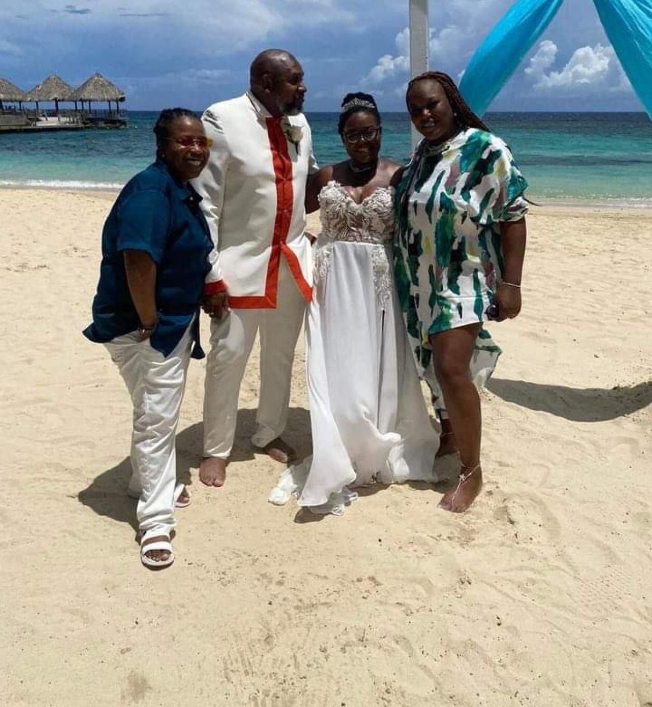 Wedding group on a beach; bride, groom, two guests. Groom in white suit; bride in white dress. Ocean in background.
