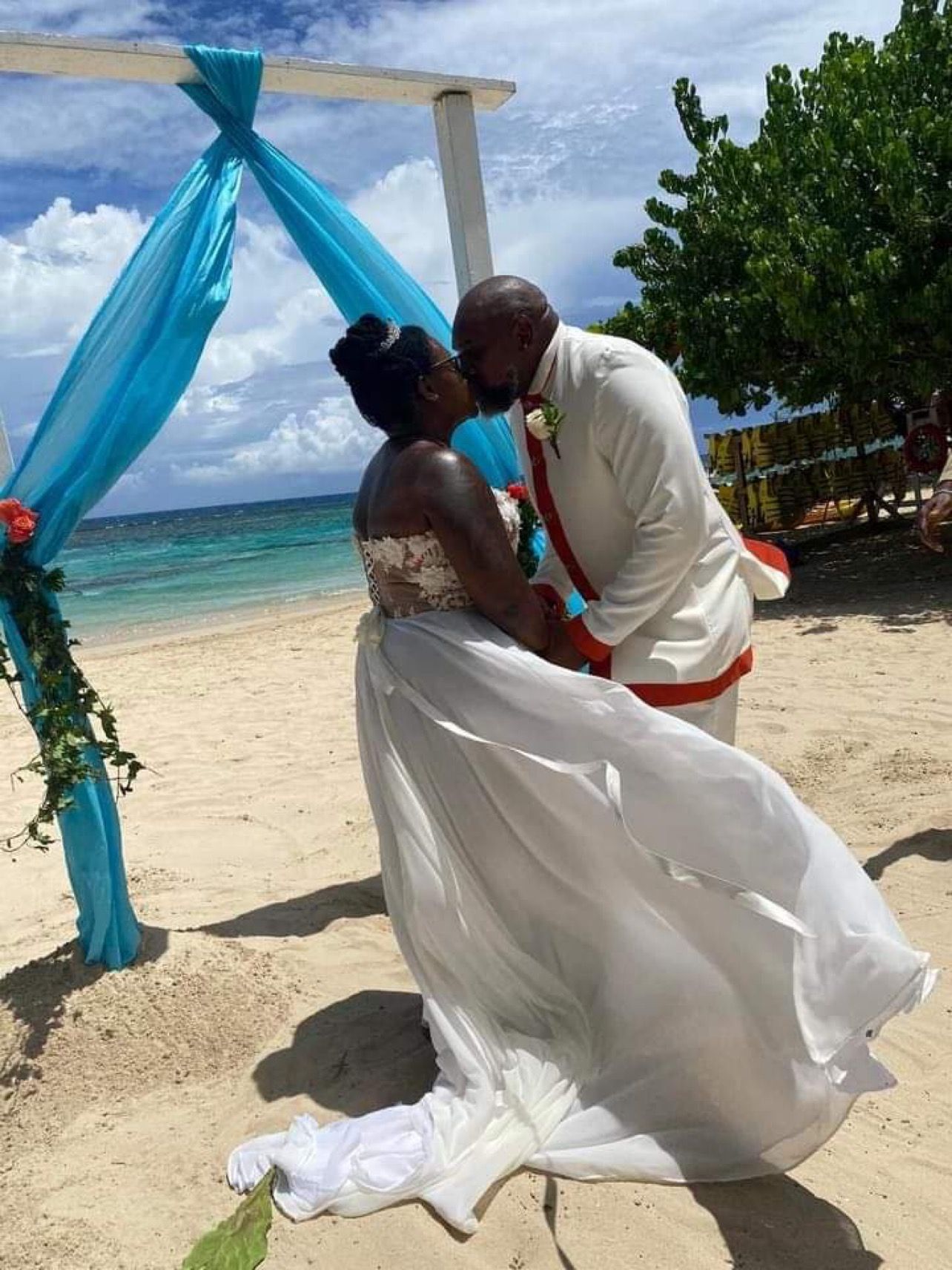 Couple kissing on a beach at their wedding. They are in wedding attire, in front of a decorated arch.
