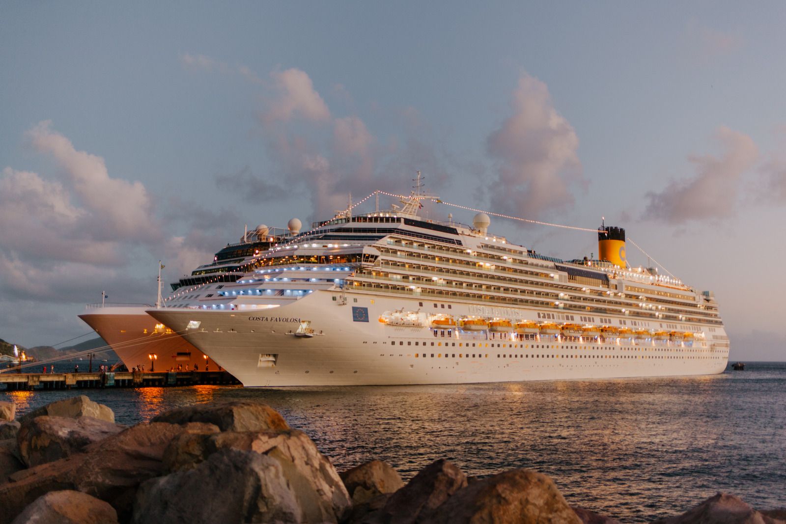 Large cruise ship docked in harbor at dusk, with lit windows and a rocky shoreline.