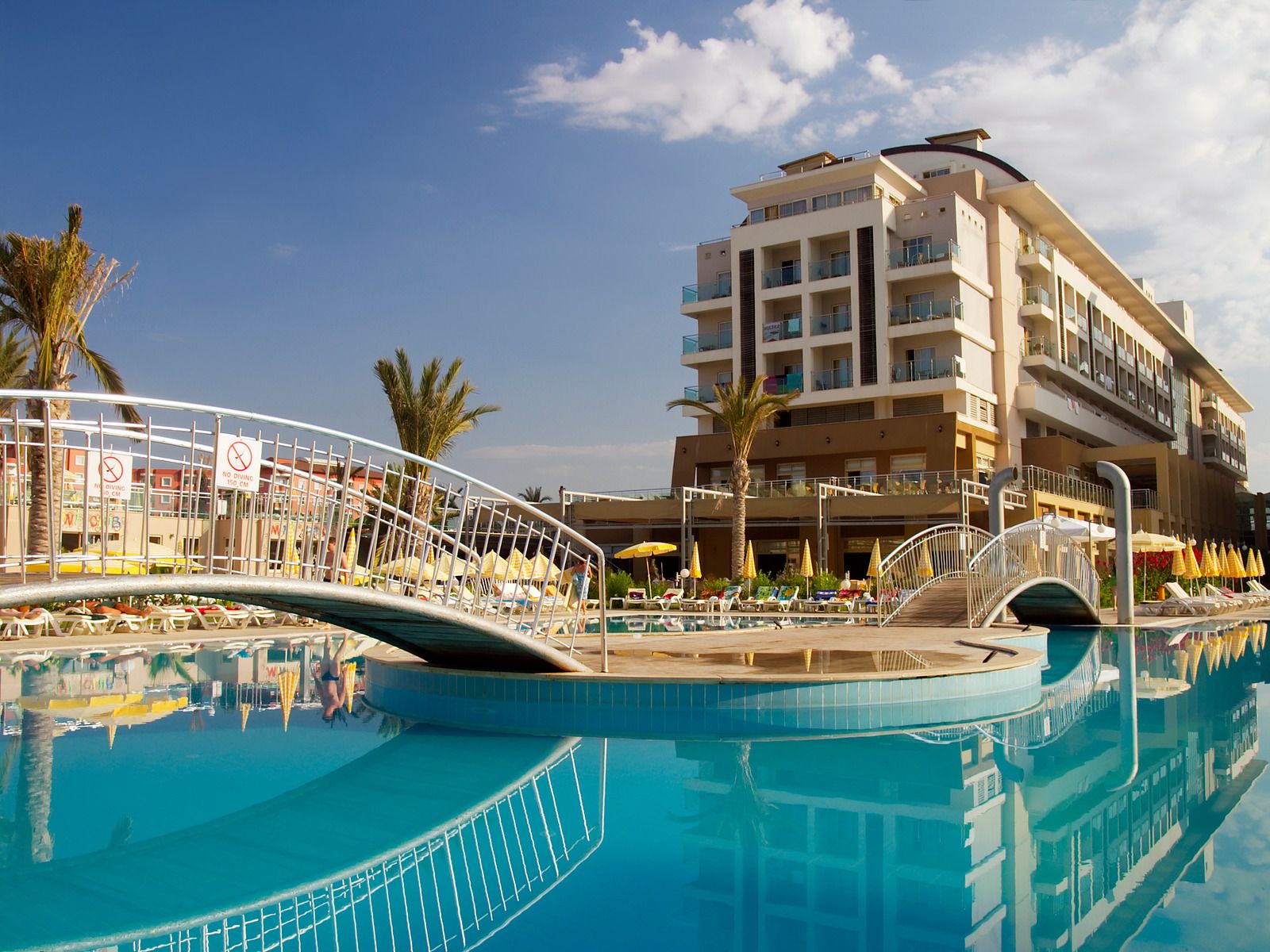 Hotel with a pool and bridge under a blue sky. Palm trees and a building are also visible.