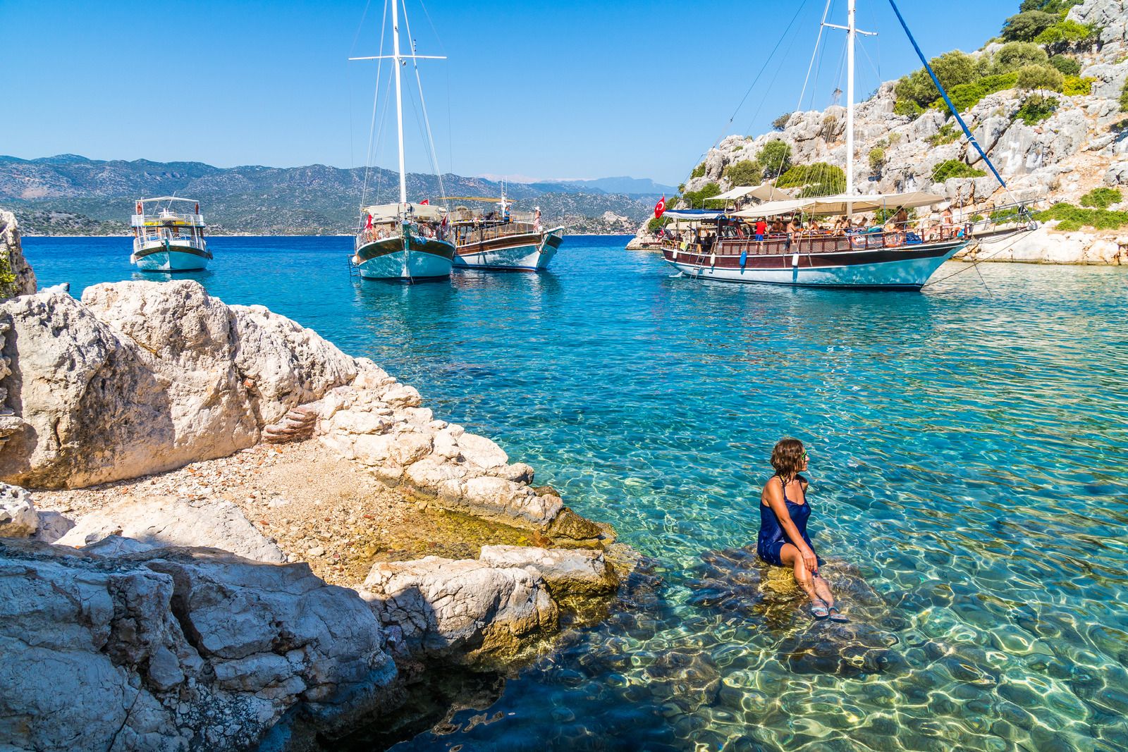 Woman sits on rocks in clear blue water, with boats in a scenic cove.
