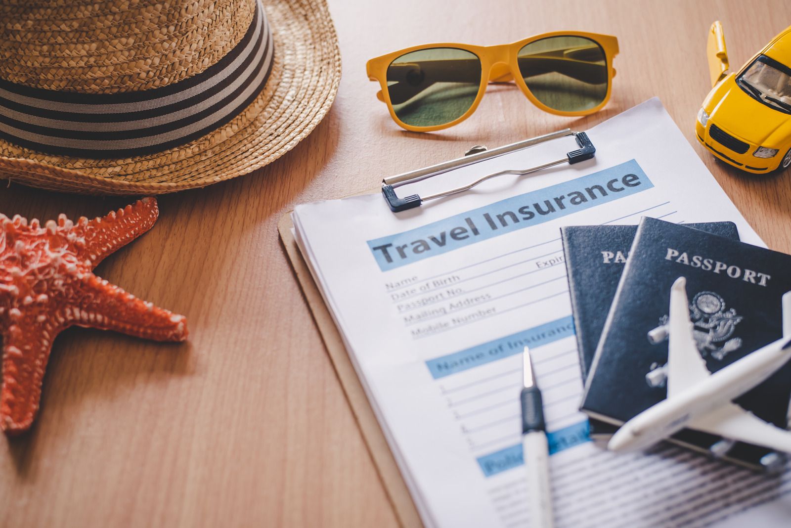 Travel essentials: hat, sunglasses, insurance form, passports, toy car, starfish, plane, pen on wooden surface.