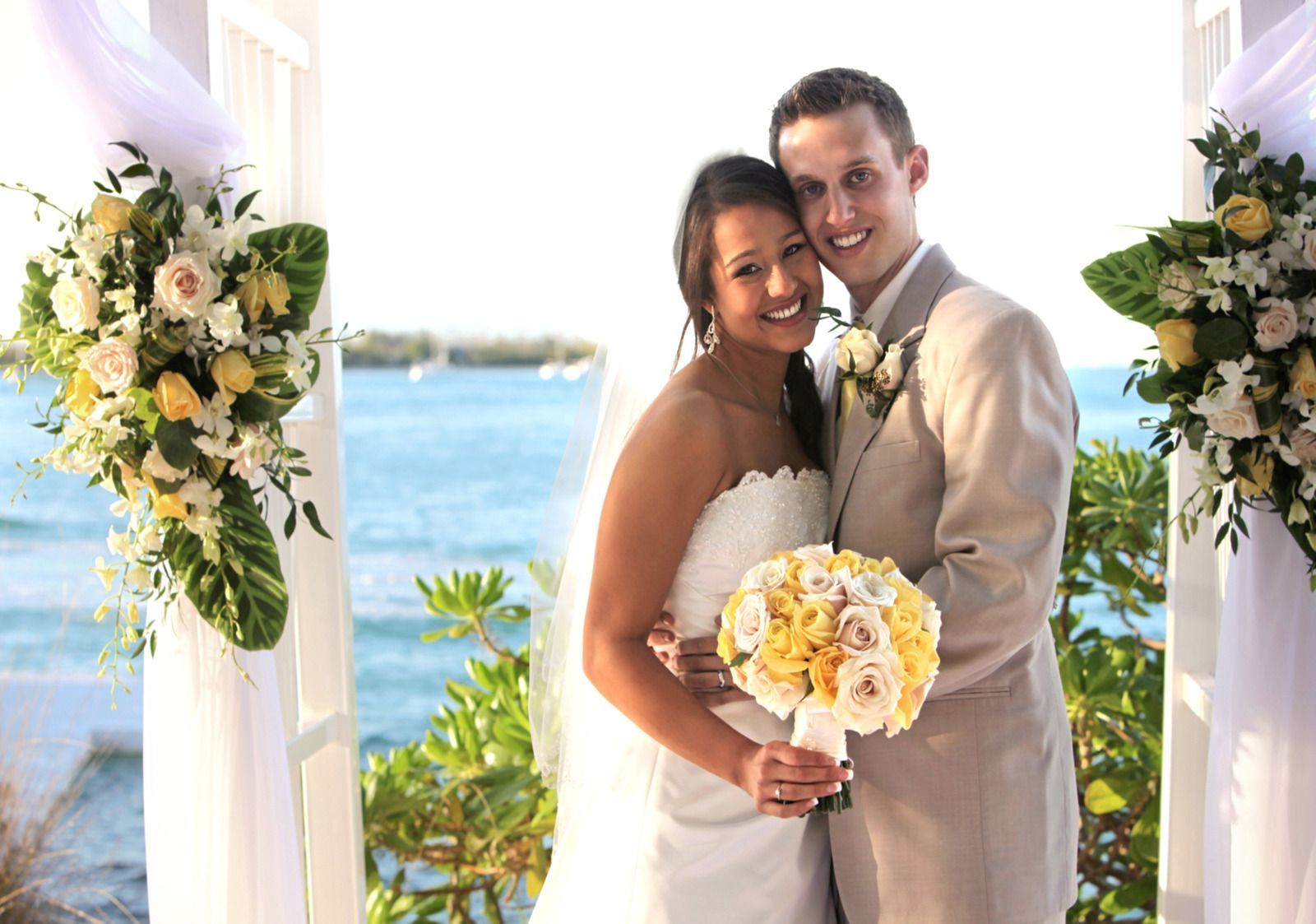 Bride and groom smiling, embracing. Wedding by the ocean, flowers and white pillars, sunny.