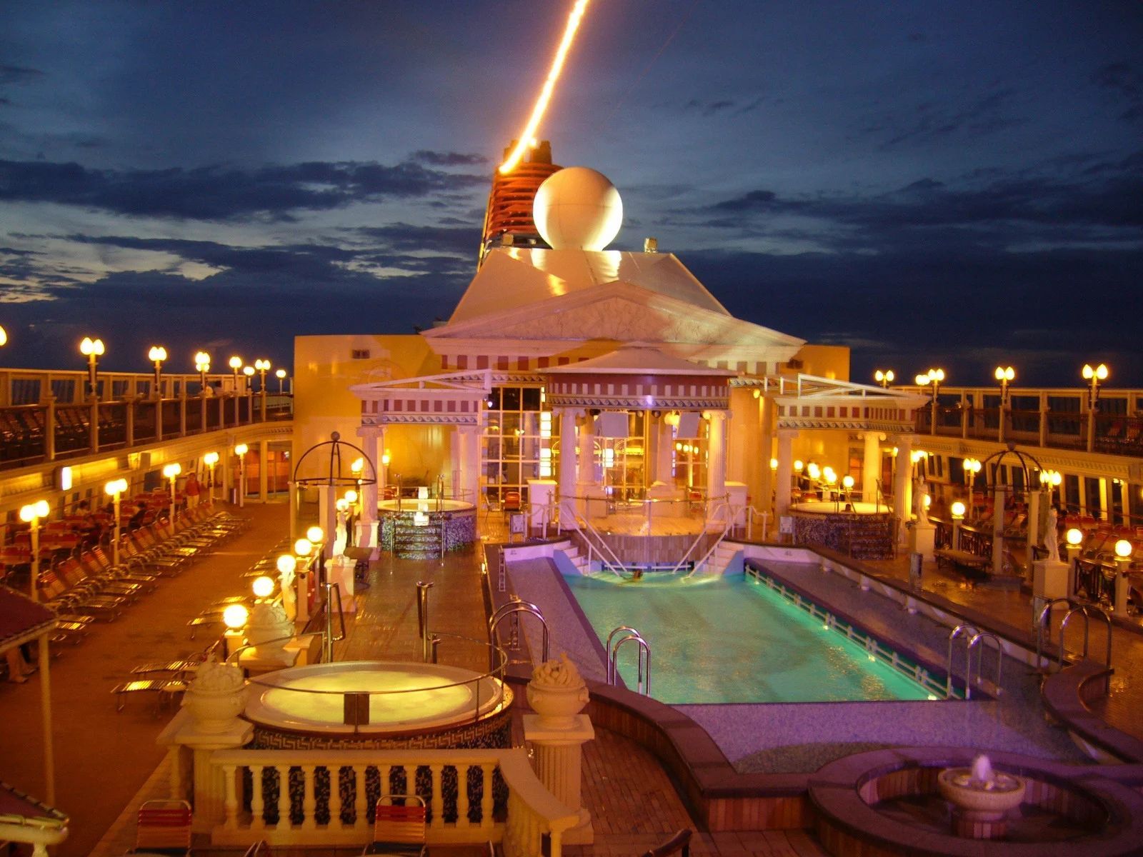 Cruise ship deck at night with lit pool, jacuzzi, and laser beam.