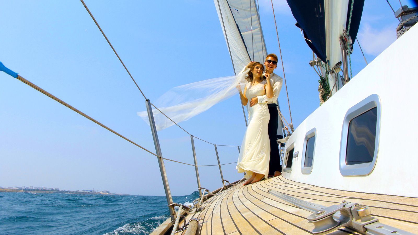 Bride and groom embrace on a sailboat deck; bride's veil blows in the wind, blue ocean, sunny day.