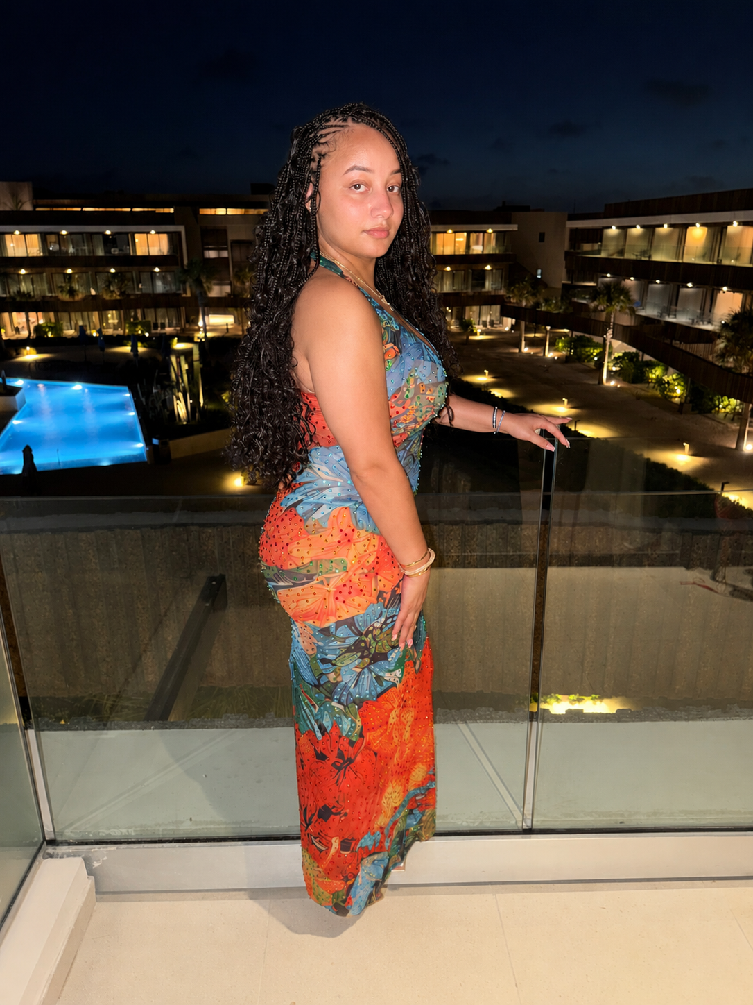 Woman in a colorful dress standing on a balcony at night, overlooking a lit pool and resort buildings.