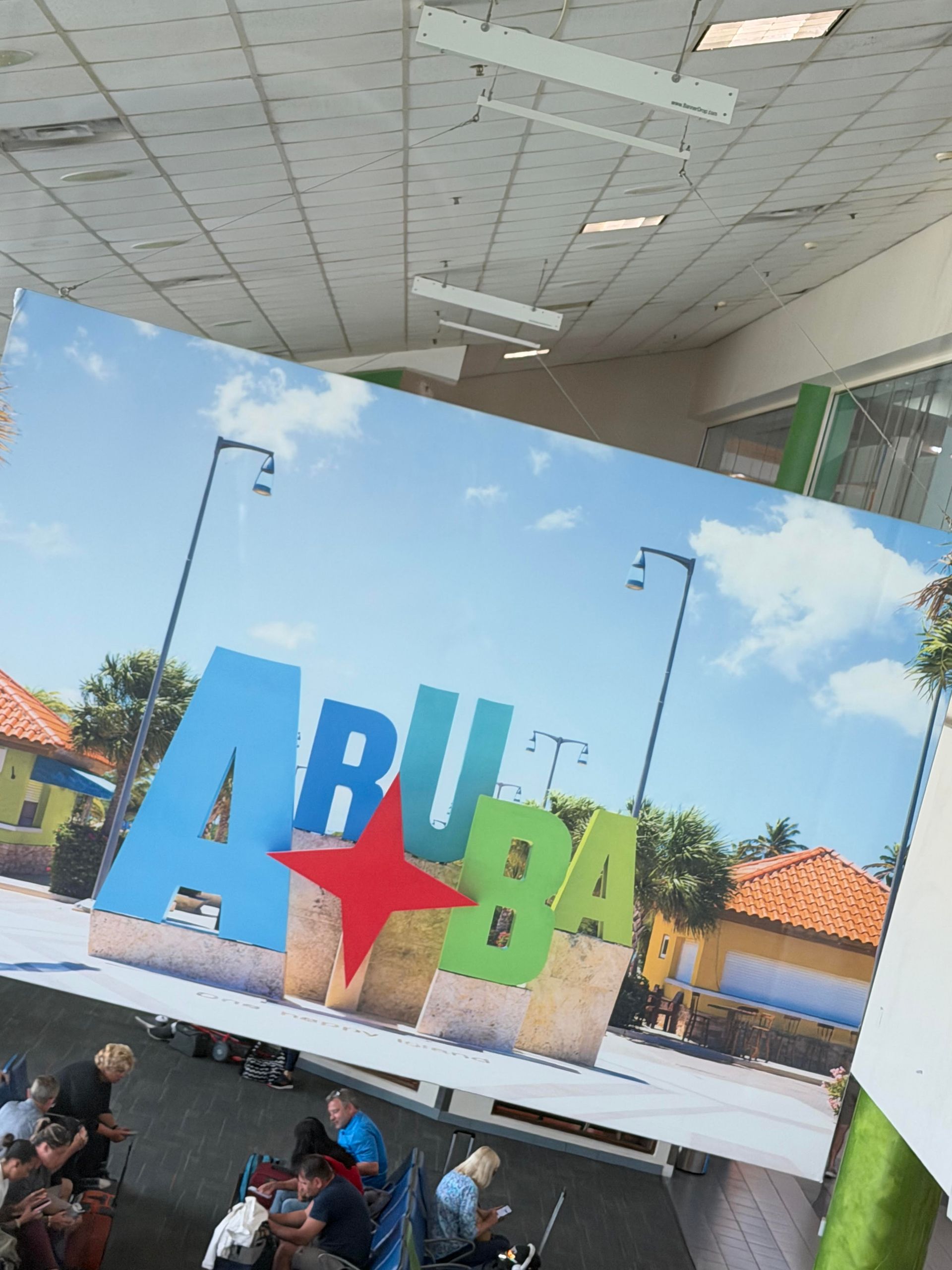 Colorful “ARUBA” sign with a red star under a cloudy sky, with people seated below in the foreground
