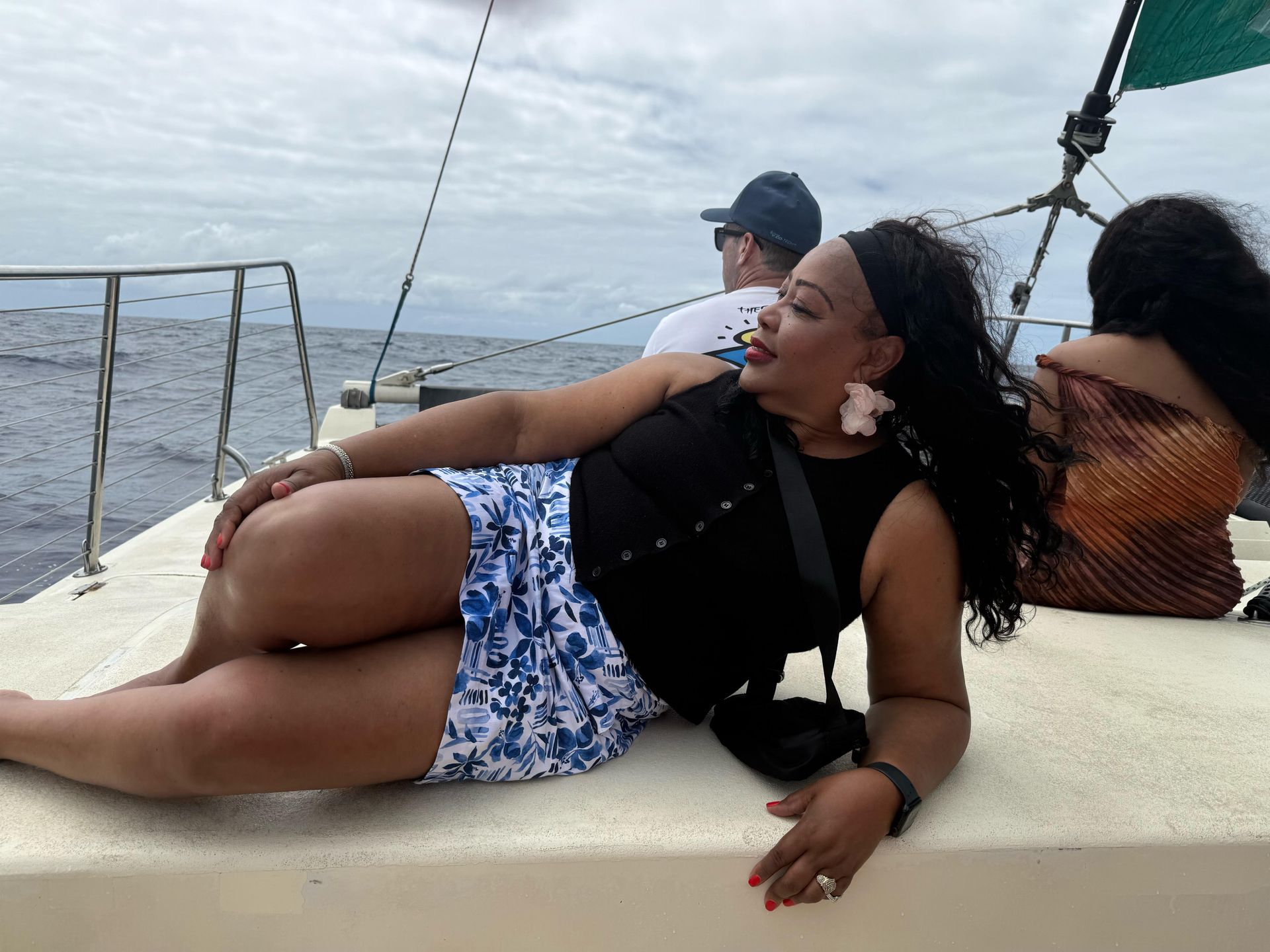 A person reclines on the deck of a boat on the water, wearing a black top and blue and white patterned shorts.