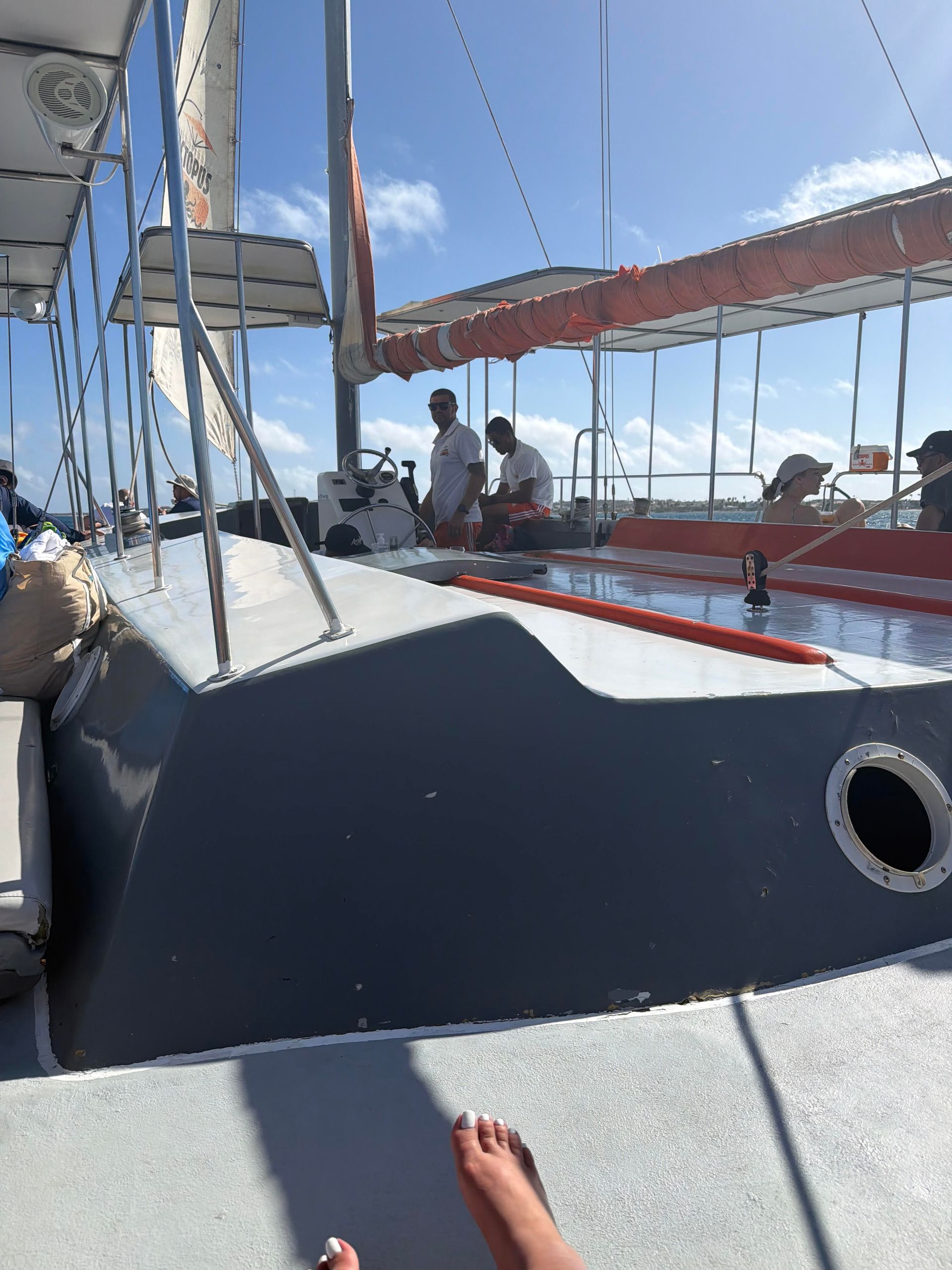 Sailboat deck with people by the mast under a clear blue sky, seen from a seated passenger's feet.