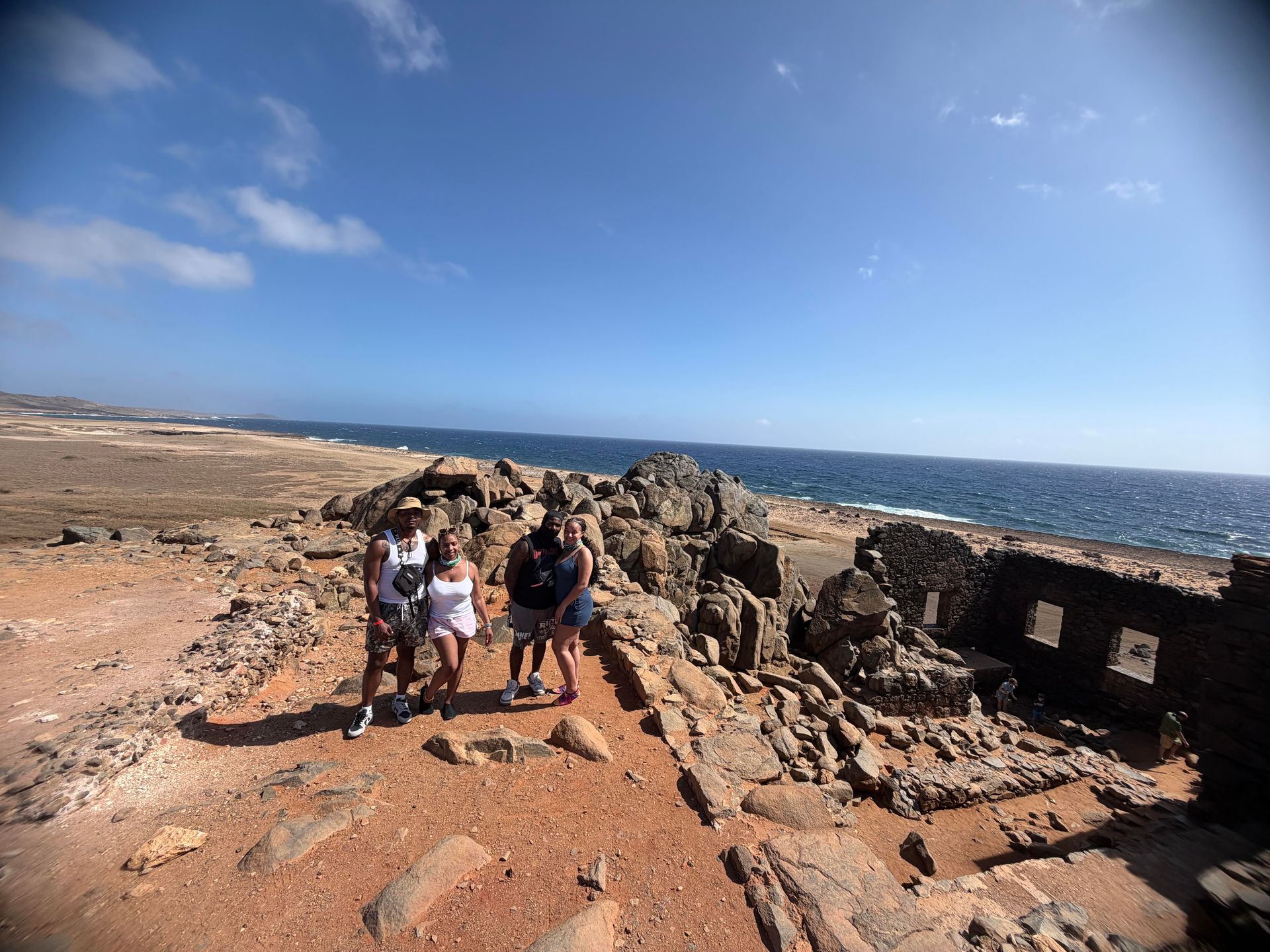People walking on a rocky coastal trail beside the sea under a bright blue sky.