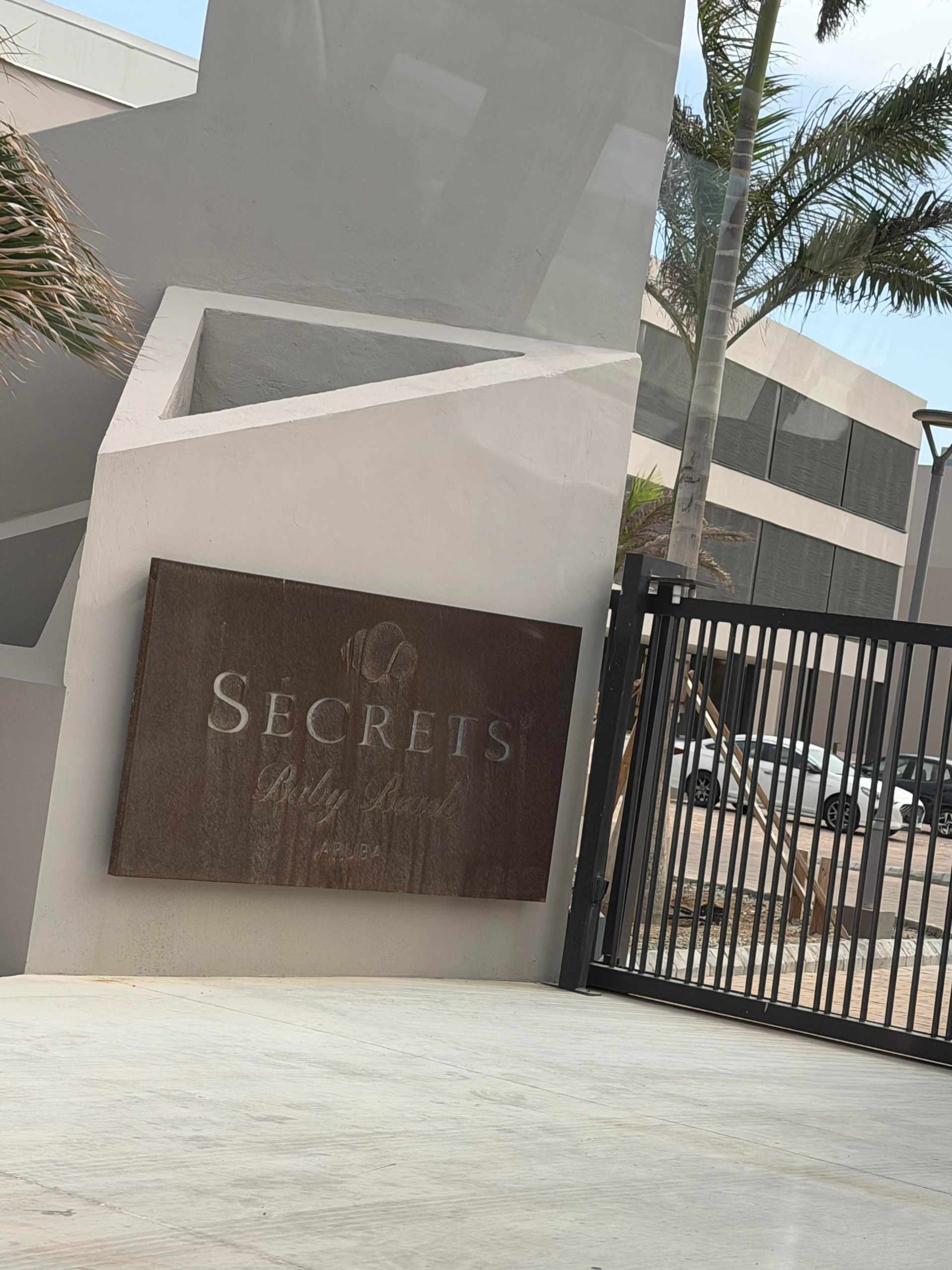 Entrance gate to Secrets resort with tan walls, metal gate, and palm trees in bright sunlight