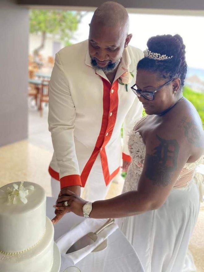 Couple cutting a wedding cake. Man in white suit with red trim, woman in white dress, outdoor setting.