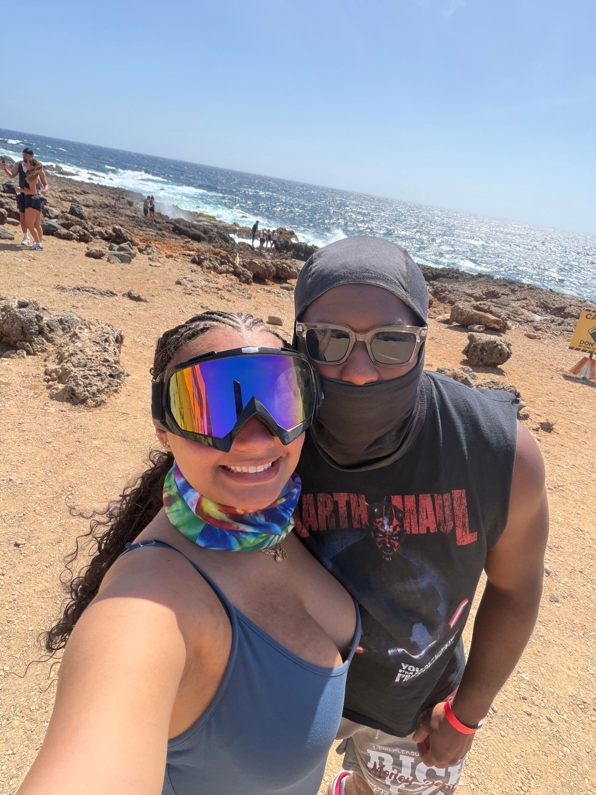 Two people taking a selfie on a rocky beach with ocean waves in the background.