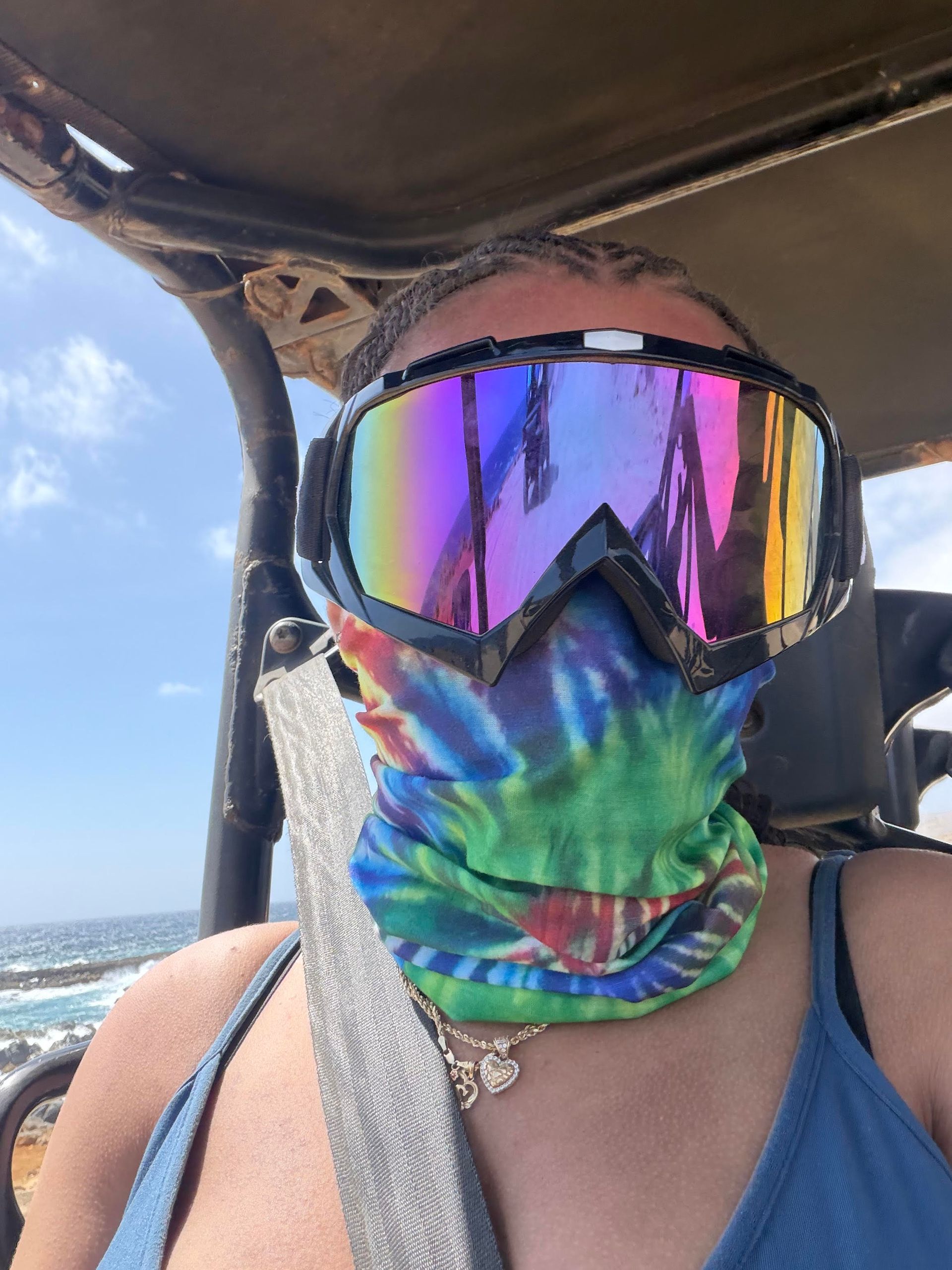 Person in mirrored ski goggles and tie-dye face covering seated on a boat with ocean in the background