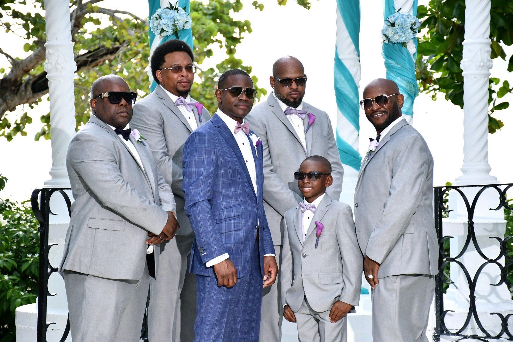 Groomsmen in gray suits and a groom in blue, posing at an outdoor wedding ceremony.