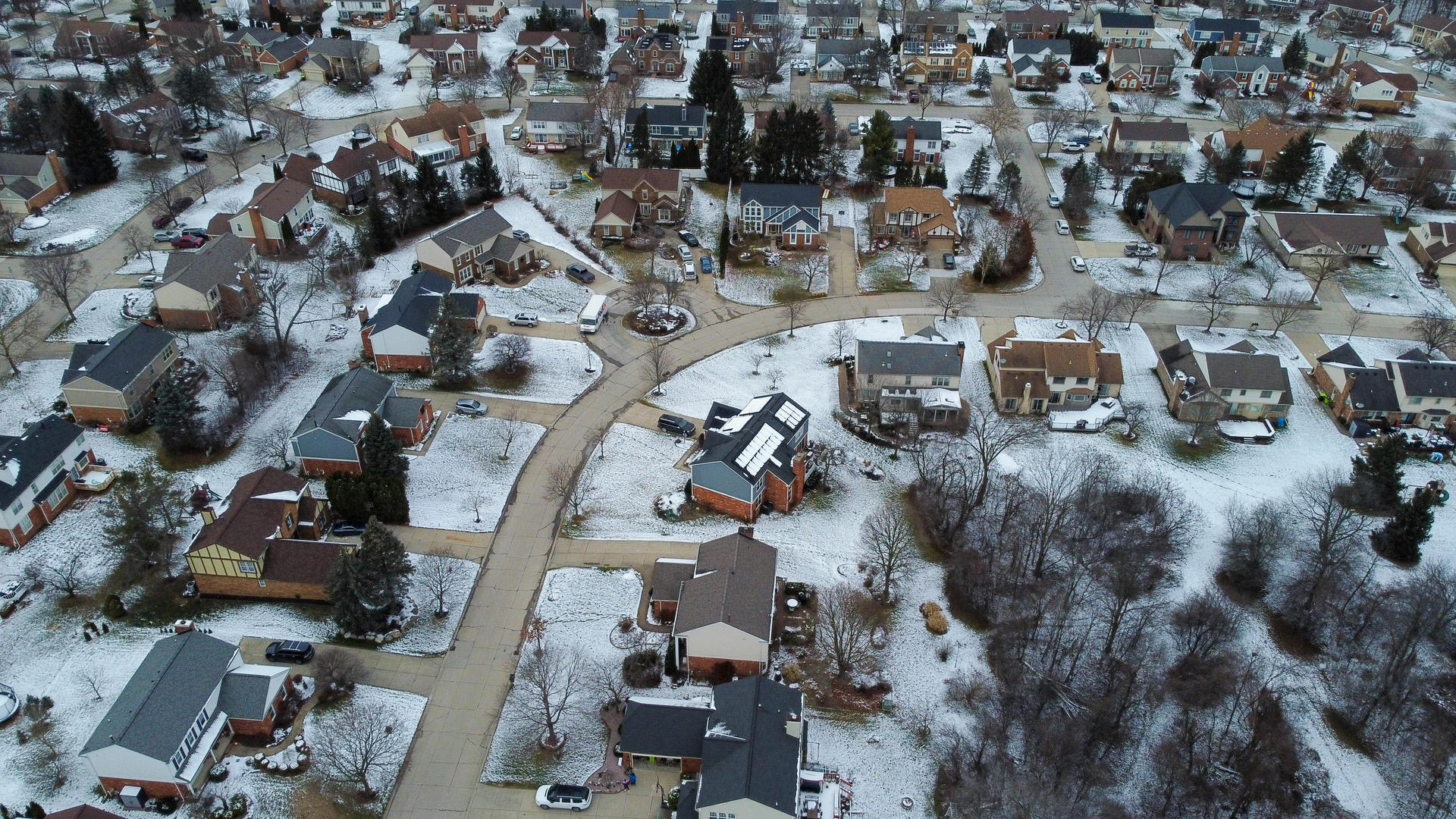 Aerial view of a suburban neighborhood covered in snow. Houses with brown and gray roofs, winding roads.