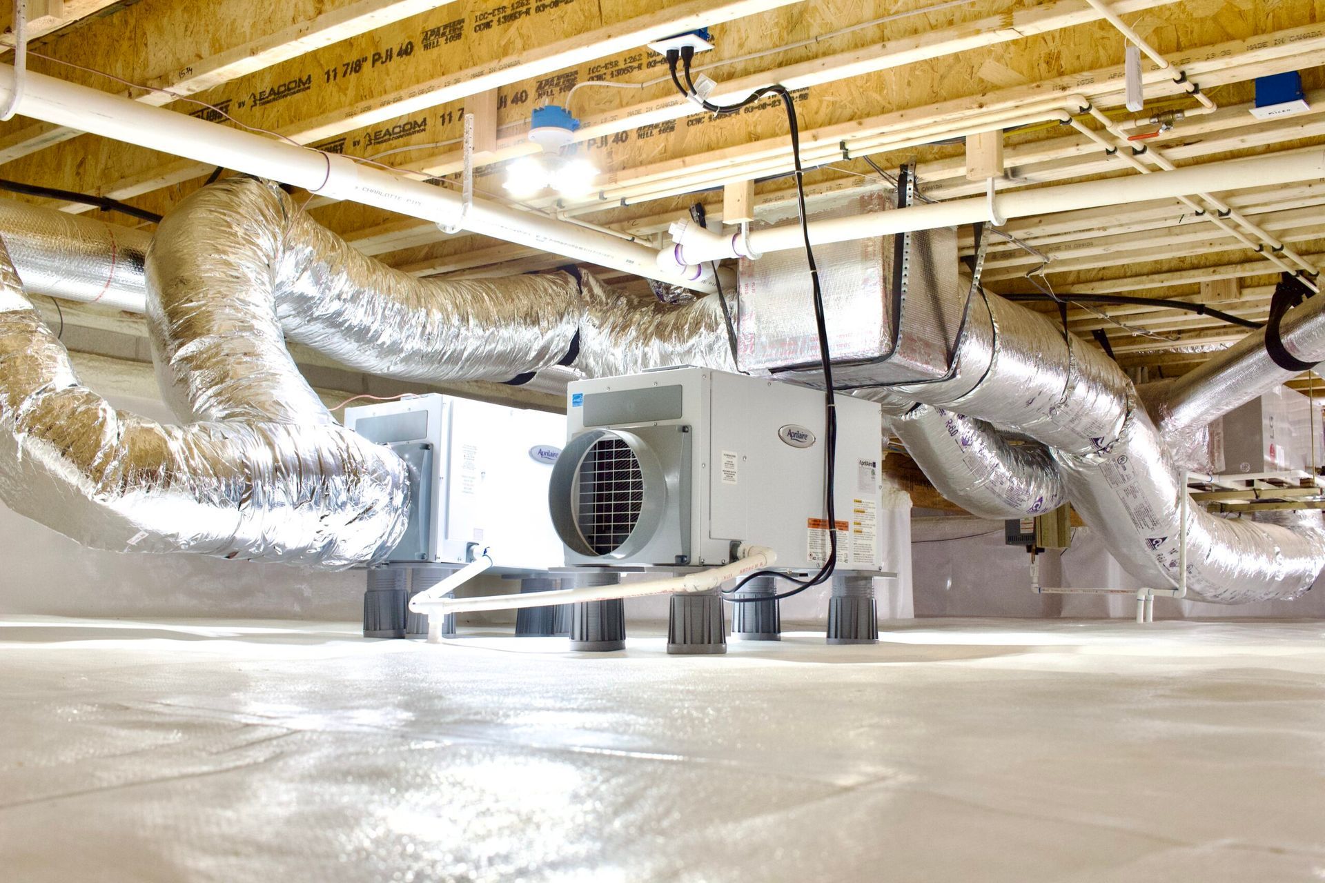 HVAC system with silver ducts in a crawl space, visible from below. White floor, wooden ceiling, and lighting.