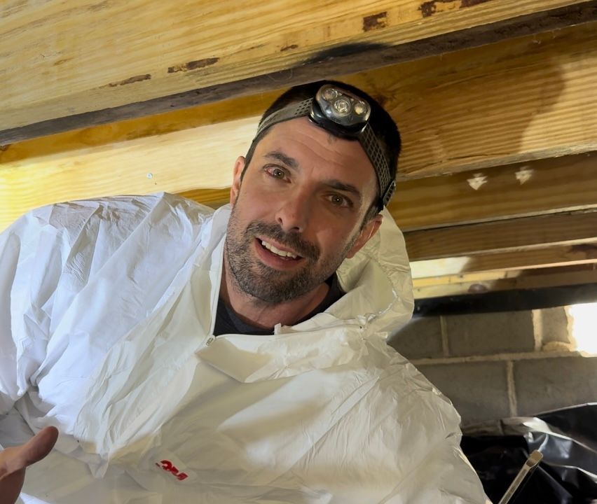 Man in a white protective suit with headlamp lying under a wooden structure, smiling at the camera.