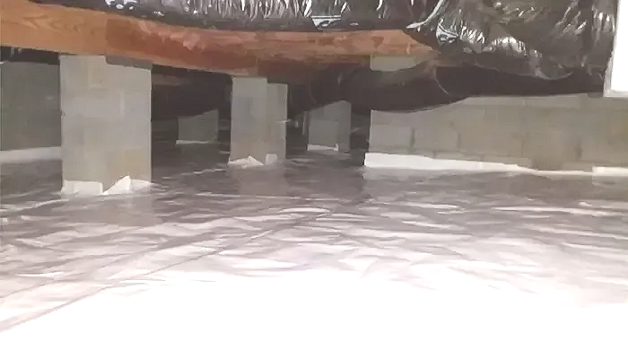 Crawlspace with silver vapor barrier on the ground, supported by concrete piers and wooden beams.