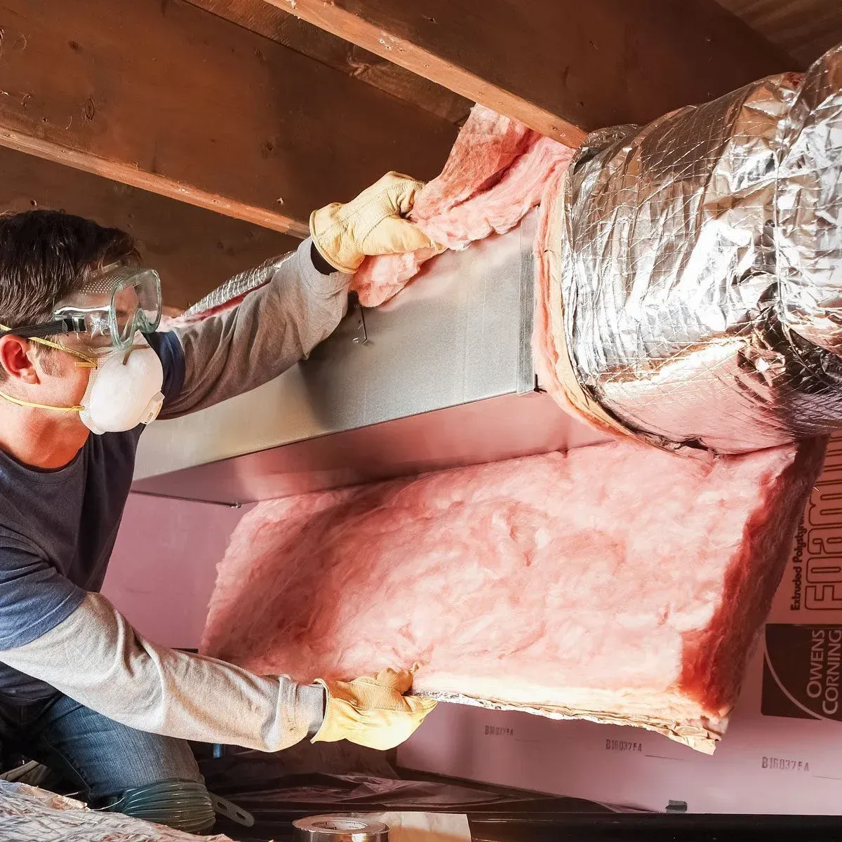 Man insulating HVAC ductwork in a crawl space, wearing safety glasses and a mask.